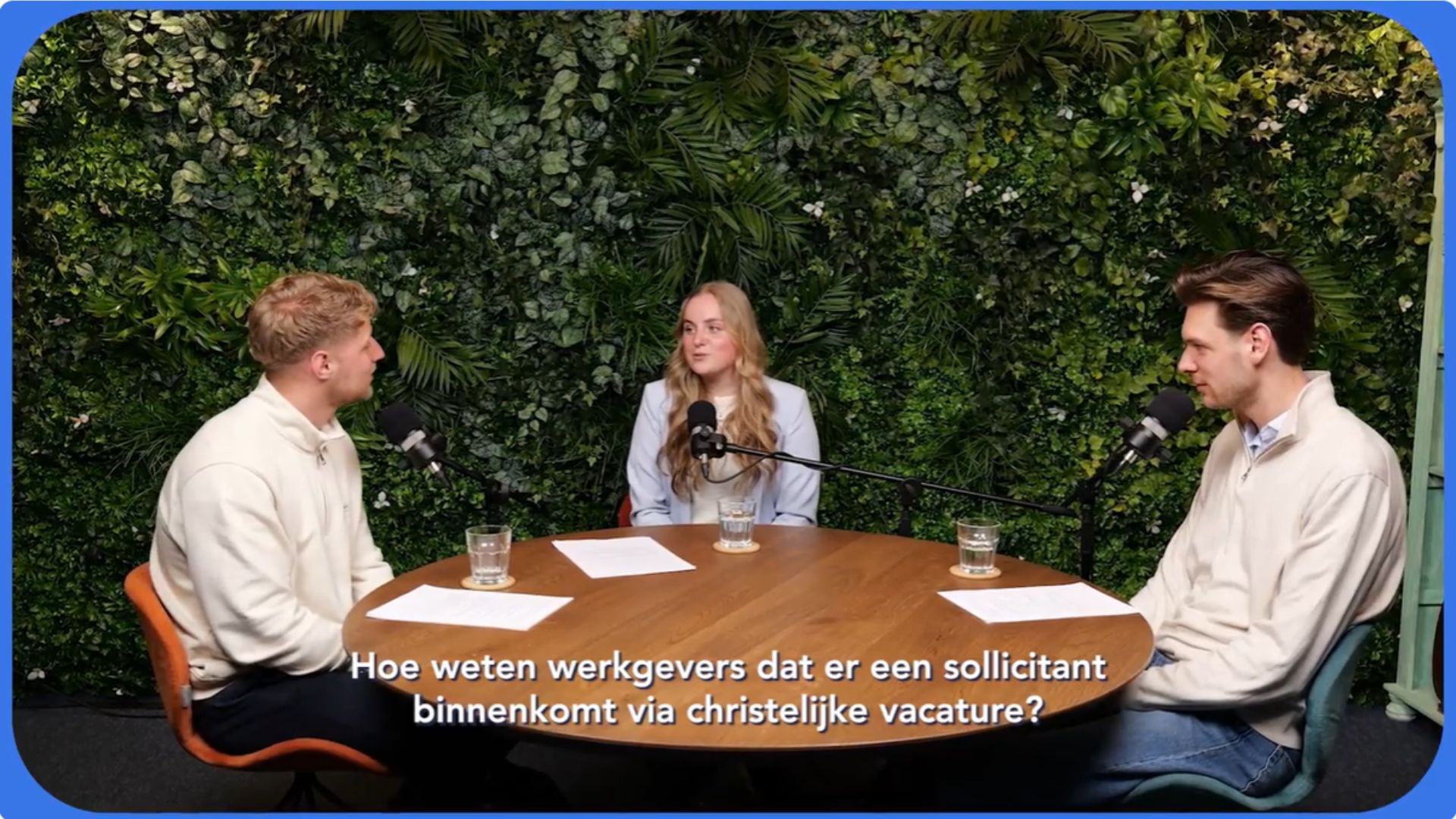 Three people seated around a wooden table with microphones, chatting in a green leafy studio backdrop (podcast/interview setting).