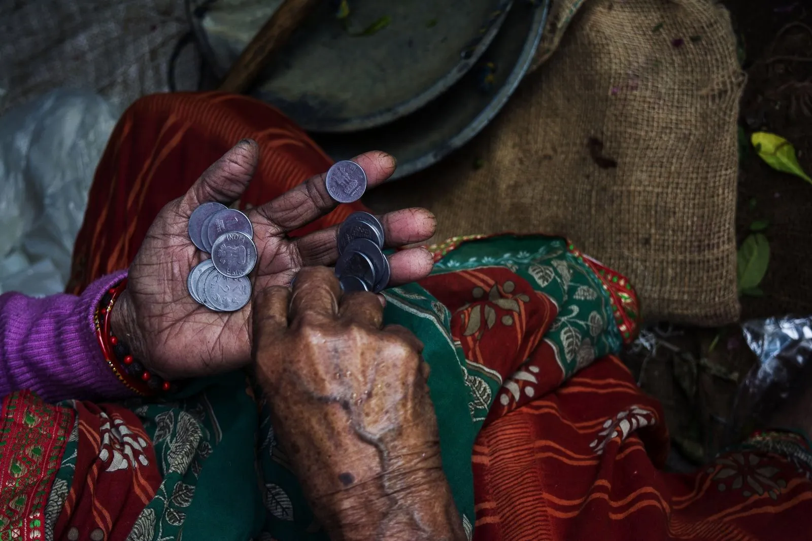 Elderly hands counting several Indian coins in a worn palm, wearing a colorful patterned sari.