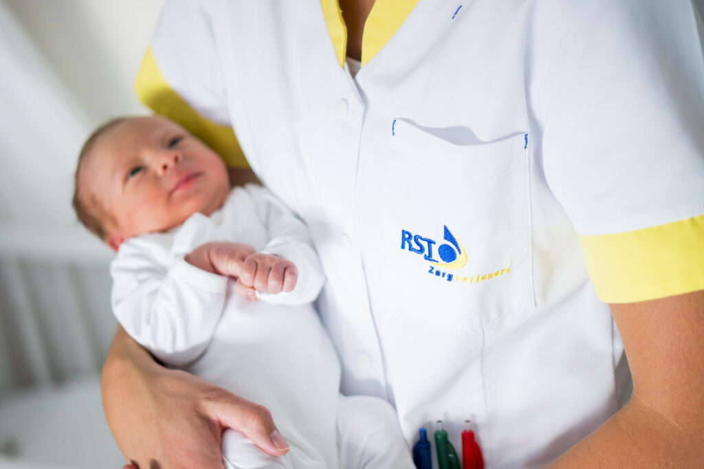 Healthcare professional in a white coat holding a newborn baby in a clinical setting, logo visible on the chest pocket.