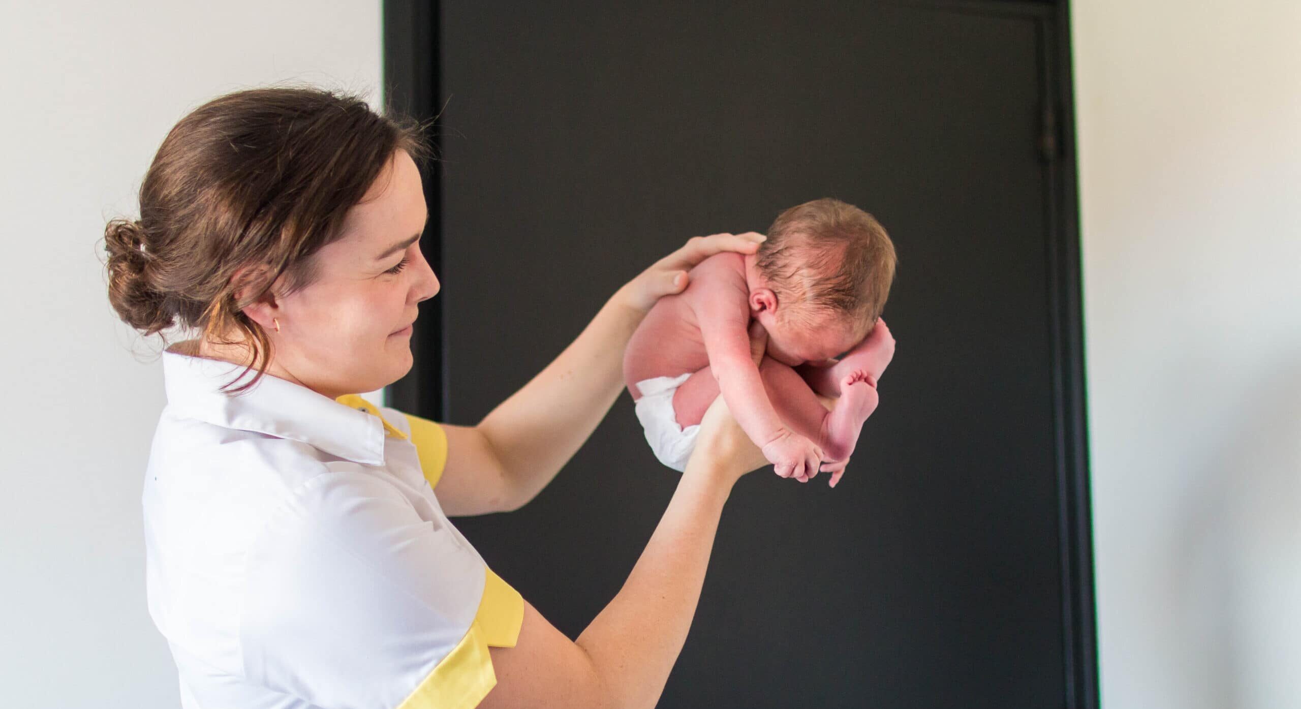 Woman in a white and yellow uniform holding a newborn baby up in the air, against a dark background on the right.