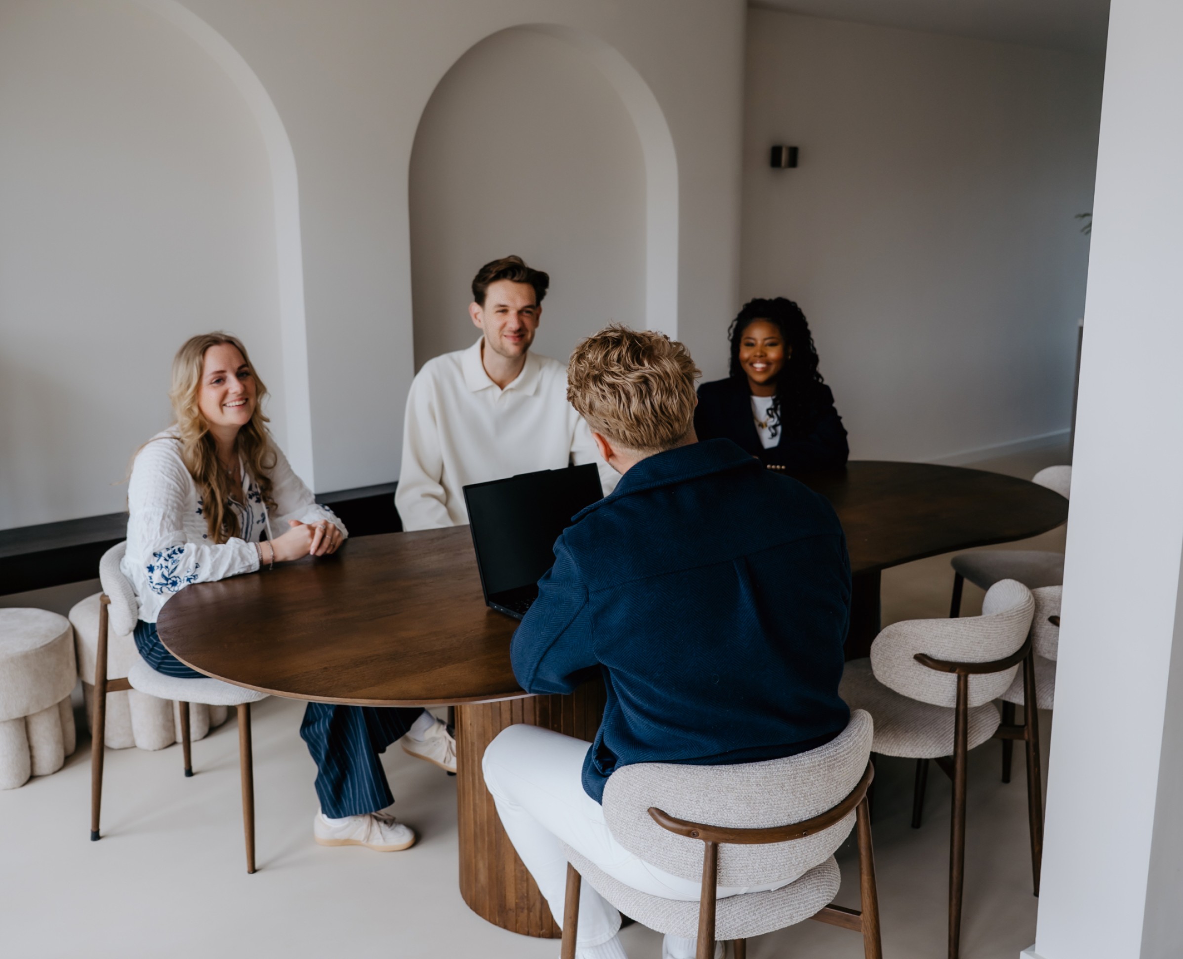 Four colleagues sit around a round wooden table in a bright, modern meeting room, smiling and talking.